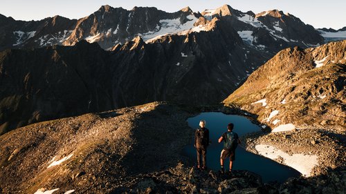 Erholungsurlaub im Chalet im Stubaital Zwei Wanderer stehen am Bergsee mit Blick auf schneebedeckte Alpenberge im Sonnenlicht